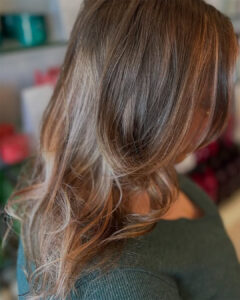 Close-up of a woman with long, wavy light brown hair—fresh from a Brazilian Blowout on Anastasia Island—wearing a dark green textured top and facing away from the camera in an indoor setting. - Get Panache, FL