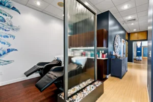 A modern St. Augustine Aveda Salon with black shampoo chairs, a glass water feature with rocks, blue feather wall art, and shelves of hair products on wood and tile floors—your destination for luxury hair extensions in St. Augustine. - Get Panache, FL