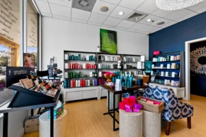 A modern salon retail area with shelves of hair and beauty products, a display table, patterned chair, chandelier, and signage for Hydrafacial Anastasia Island and highly-skilled lash extensions St. Augustine. - Get Panache, FL