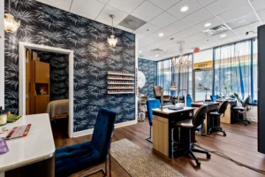 Modern nail salon interior with manicure stations, blue-patterned accent wall, nail polish display, blue chairs, and natural light from large windows—perfectly complementing the style of a St. Augustine Aveda Salon. - Get Panache, FL