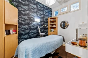 A treatment room featuring a bed with a blue quilt, wooden cabinets, a wall-mounted lamp, skincare products, and a decorative mirror against dark botanical wallpaper—perfect for your Hydrafacial Anastasia Island or luxury deep tissue massage St. Augustine. - Get Panache, FL