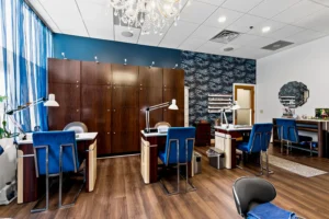 Modern nail salon interior at a St. Augustine Aveda Salon, featuring blue chairs, wooden manicure tables, wall-mounted polish racks, and a decorative chandelier on a wood floor. - Get Panache, FL