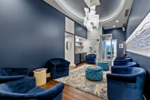 A modern waiting room with navy blue walls, blue velvet chairs, a textured rug, round ottomans, wall art, and a contemporary light fixture welcomes guests seeking luxury hair extensions or highly-skilled lash extensions in St. Augustine. - Get Panache, FL