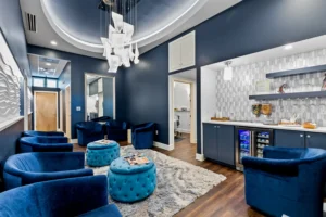 Modern waiting room with blue velvet chairs, round ottomans, a rug, wall art, a snack station, and overhead contemporary lighting—perfect for relaxing before your highly-skilled lash extensions St. Augustine or luxury deep tissue massage St. Augustine. - Get Panache, FL