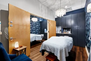 A massage therapy room with a treatment table, blue patterned walls, a chandelier, cabinets, and a side table with a salt lamp—perfect relaxation before your Hydrafacial Anastasia Island appointment. - Get Panache, FL