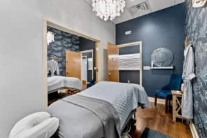 A spa treatment room at a St. Augustine Aveda Salon, featuring a massage table, blue patterned wallpaper, a chair, mirror, and an open door leading to more luxury services like highly-skilled lash extensions in St. Augustine. - Get Panache, FL
