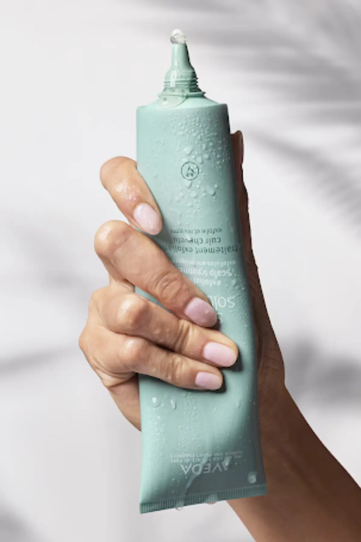 A hand with manicured nails holds a teal tube of Aveda product with water droplets on it; a small amount of product for frizz control for humidity in St. Augustine is visible at the nozzle. - Get Panache, FL
