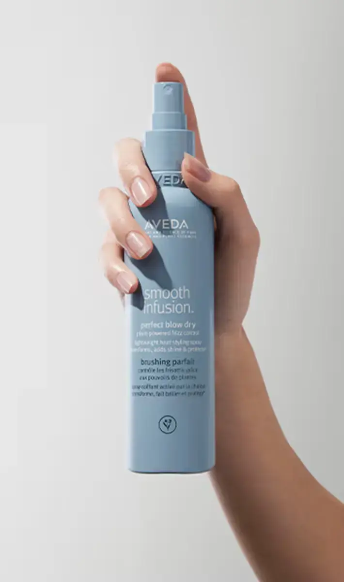 A hand holding a blue bottle of Aveda Smooth Infusion Perfect Blow Dry spray against a plain white background, reflecting the essence of self-care and wellness in St. Augustine. - Get Panache, FL