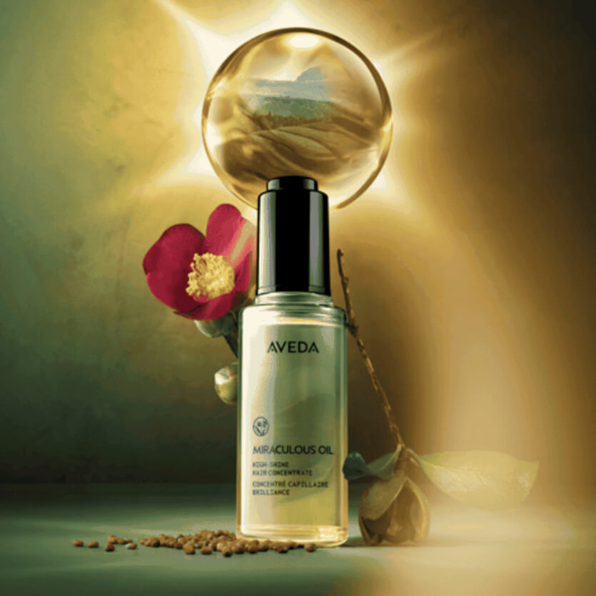 A clear bottle of Aveda Miraculous Oil is placed in front of a glowing orb, with a red camellia flower and scattered seeds nearby. - Get Panache, FL