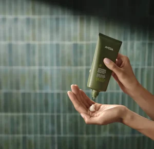 A person squeezes a small amount of Aveda Botanical Repair conditioner from a green tube onto their palm, featuring the nourishing power of Aveda Advanced Botanical Kinetics, against a tiled background. - Get Panache, FL