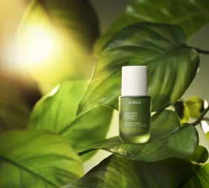 A green bottle of Aveda Advanced Botanical Kinetics Vitality Serum rests on lush leaves, illuminated by sunlight in the background. - Get Panache, FL