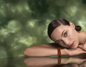 A woman with dark hair in a low bun rests her head and arms on a reflective surface, wearing a red beaded bracelet. The serene scene, paired with the green blurred background, evokes the calm of Aveda Advanced Botanical Kinetics skincare. - Get Panache, FL