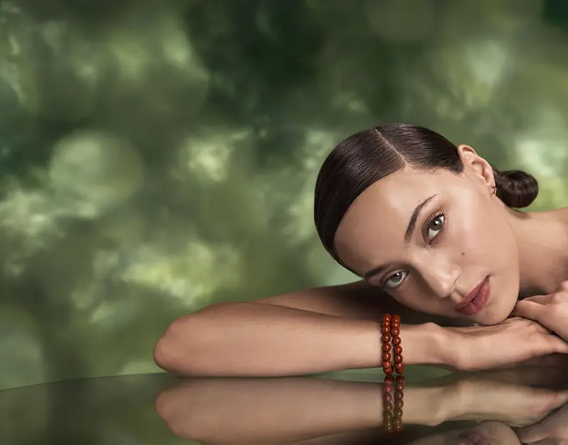 A woman with dark hair in a low bun rests her head and arms on a reflective surface, wearing a red beaded bracelet. The serene scene, paired with the green blurred background, evokes the calm of Aveda Advanced Botanical Kinetics skincare. - Get Panache, FL