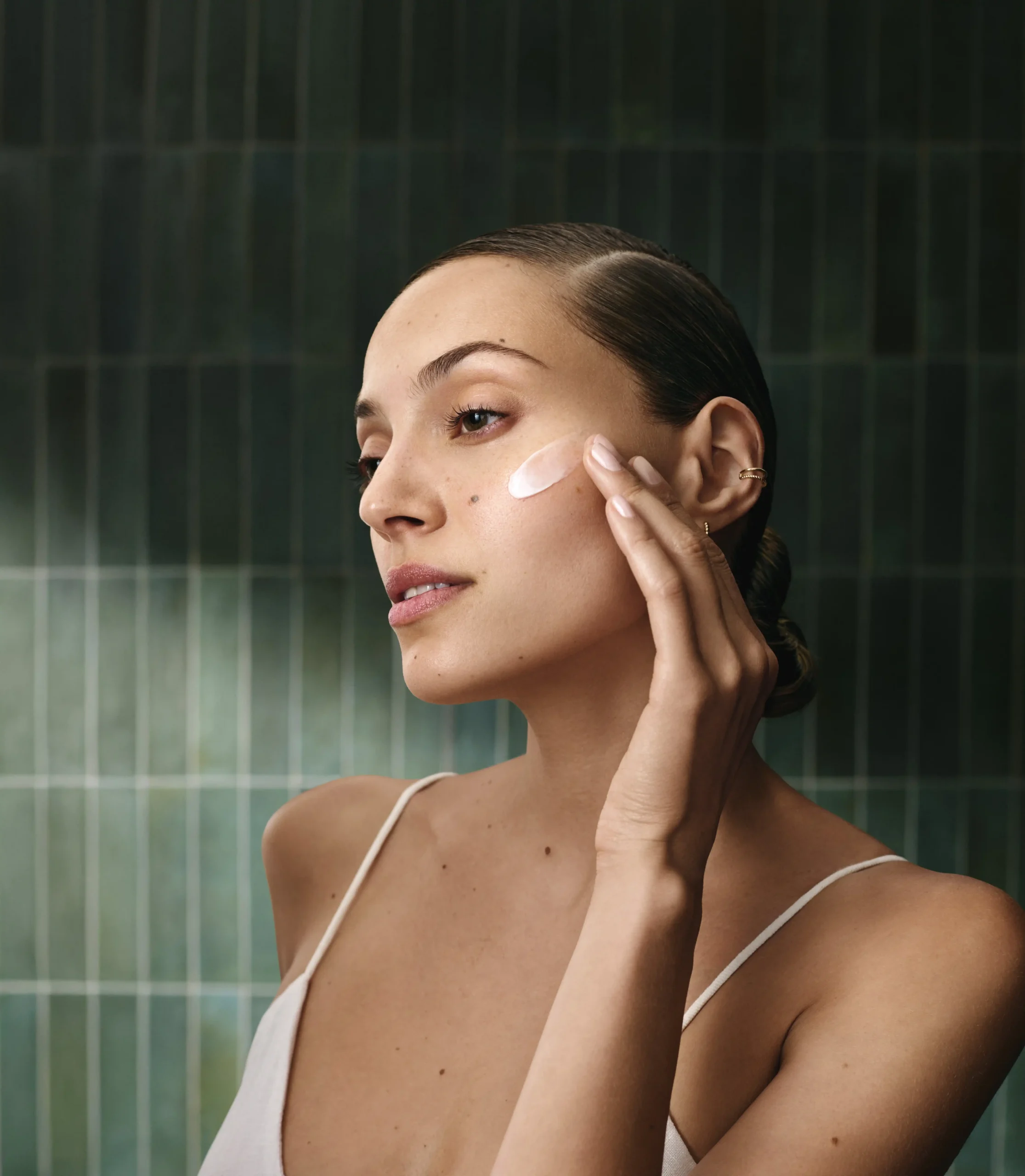 A woman applies Aveda Advanced Botanical Kinetics cream to her cheek with her fingers, standing in front of a green tiled wall. - Get Panache, FL