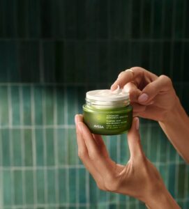 A person holds an open jar of Aveda Advanced Botanical Repair Intensive Mask, with a small amount of cream on their finger. The green tiled background reflects the clean, nature-inspired feel of the Aveda Advanced Botanical Kinetics line. - Get Panache, FL