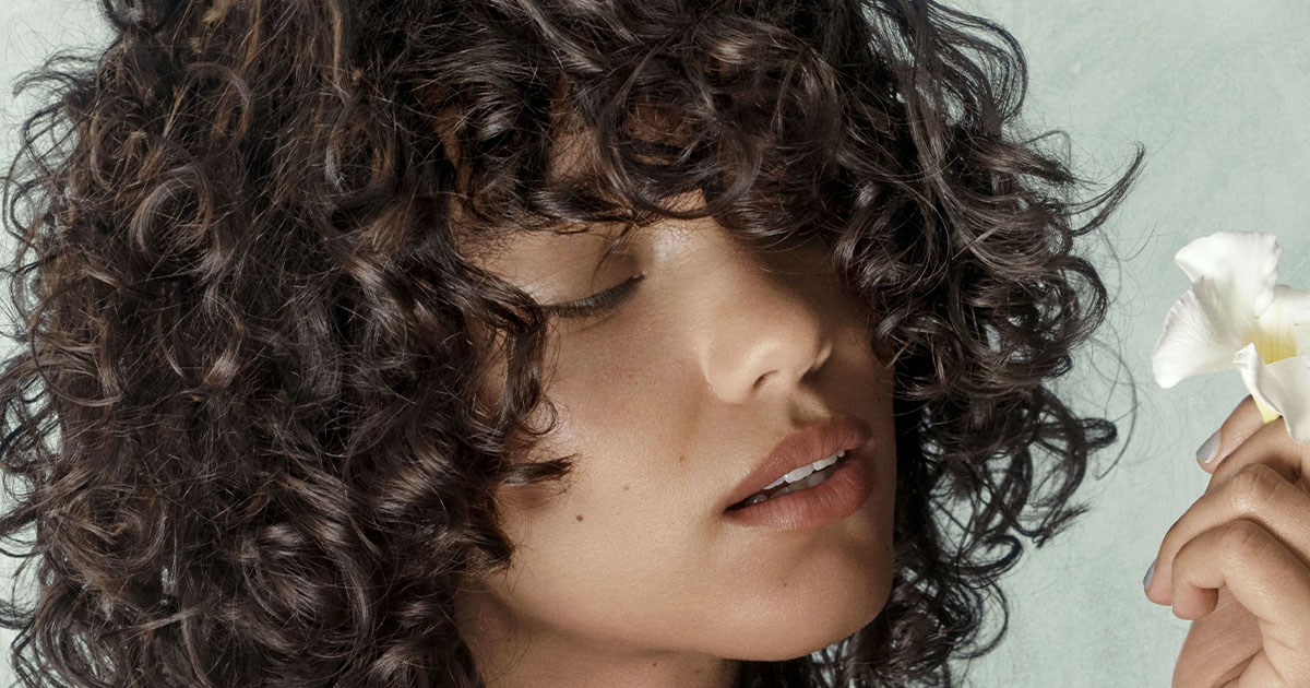 A person with curly dark hair holds a white flower close to their face, eyes closed, against a light background. - Get Panache, FL
