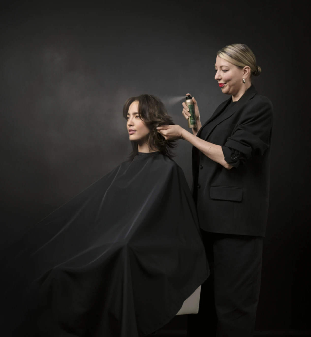 A hairstylist sprays product onto a seated client's hair in a studio with a black background. - Get Panache, FL