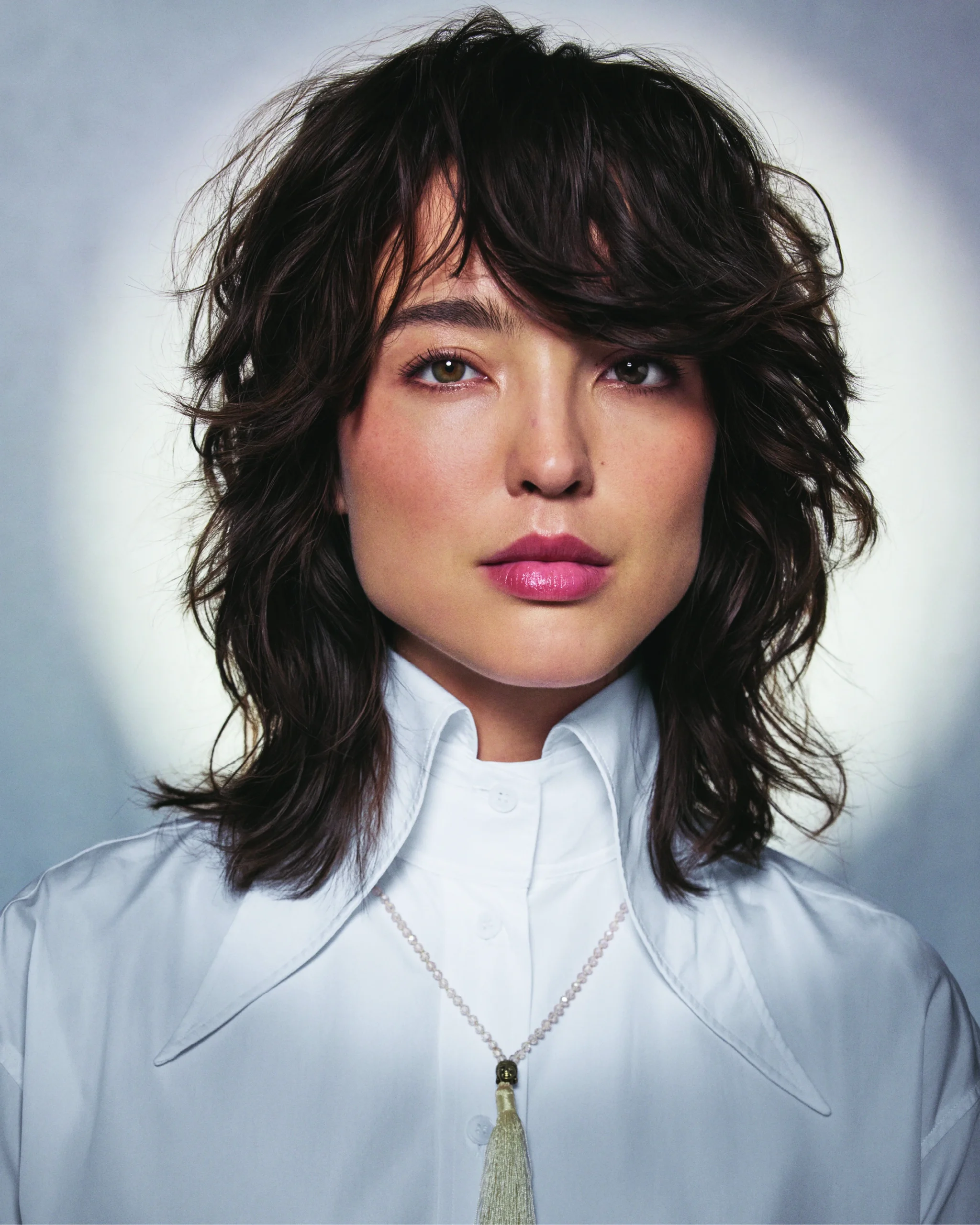 Person with wavy, layered brown hair wearing a white collared shirt and a necklace, posed against a neutral background with a circular light effect. - Get Panache, FL