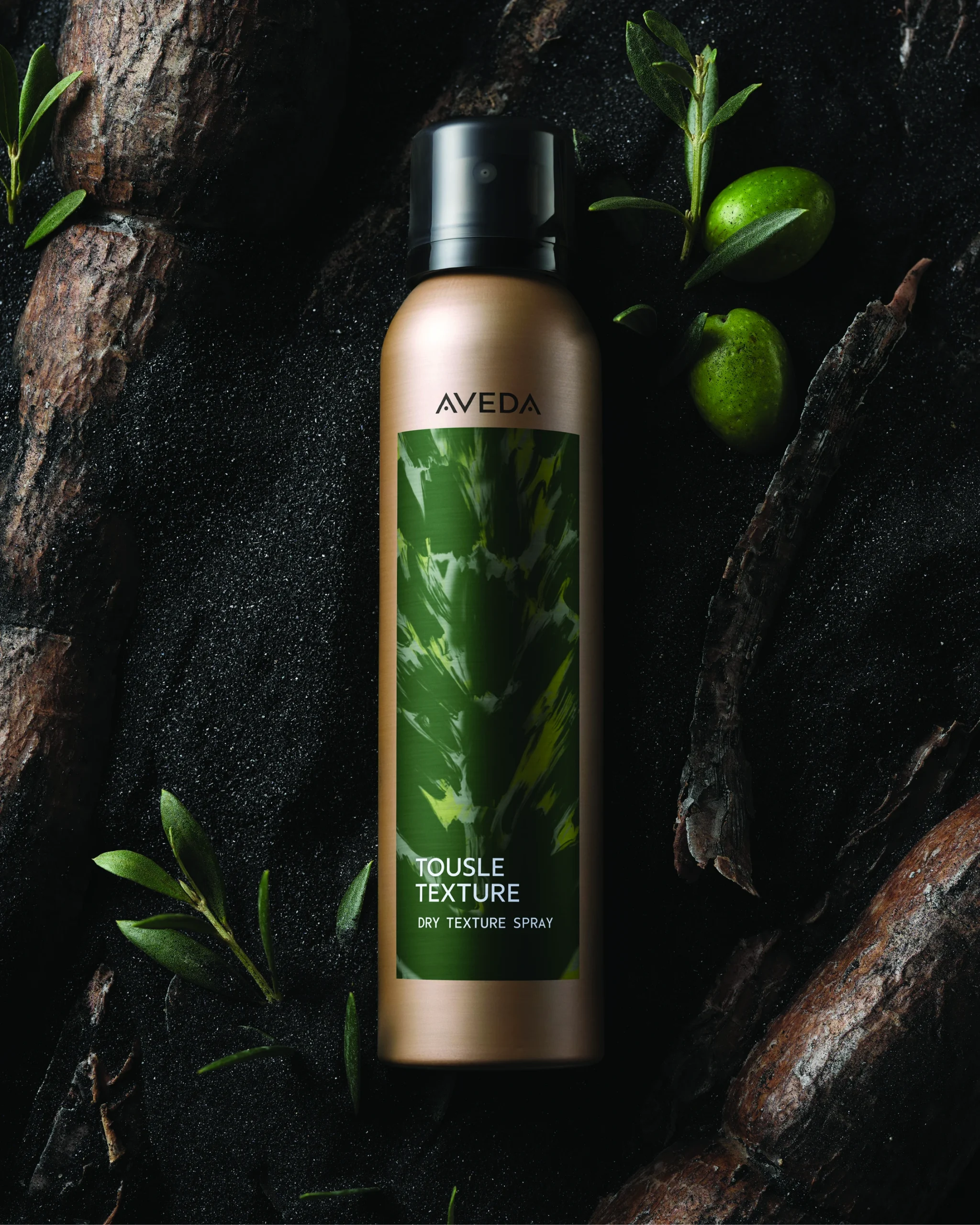 Aveda Tousle Texture Dry Texture Spray bottle on a dark surface with green leaves and small green fruits nearby. - Get Panache, FL