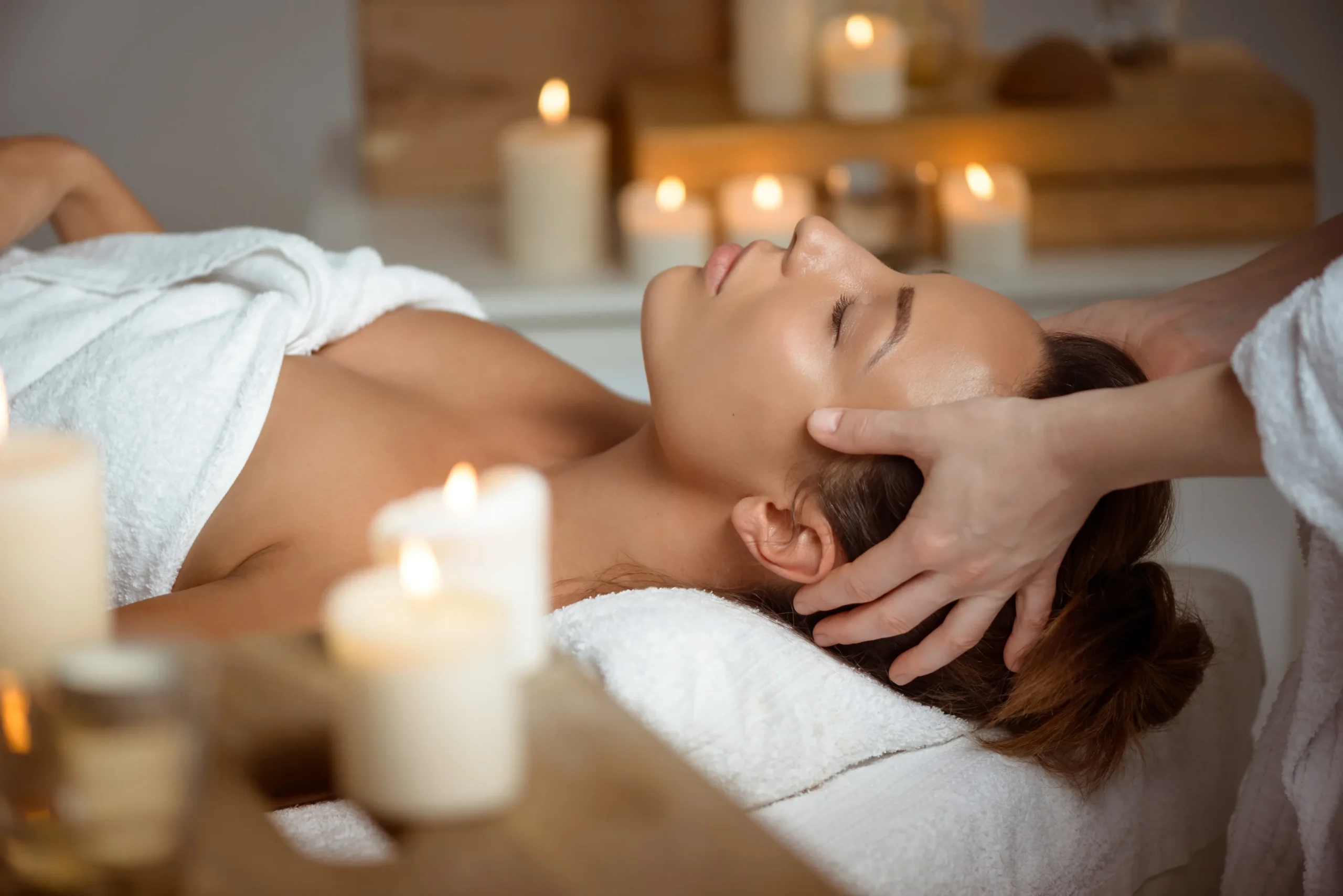 Person lying on a massage table with eyes closed, receiving a head massage, surrounded by lit candles and soft lighting in a spa setting. - Get Panache, FL