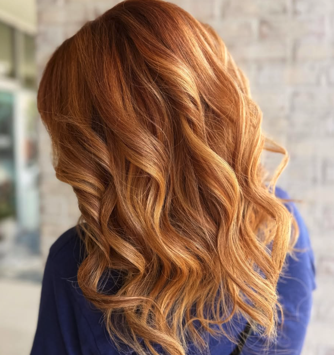 Person with long, wavy, copper-toned hair facing away from the camera, wearing a blue top, standing in front of a blurred brick wall background. - Get Panache, FL