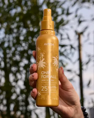 A hand holds a gold spray bottle of Aveda One For All 25 Benefits hair product outdoors, with green plants and a blue sky in the background. - Get Panache, FL