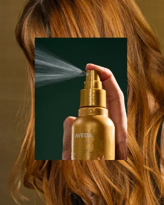 A person sprays a gold bottle of Aveda product in front of long, wavy, light brown hair. - Get Panache, FL